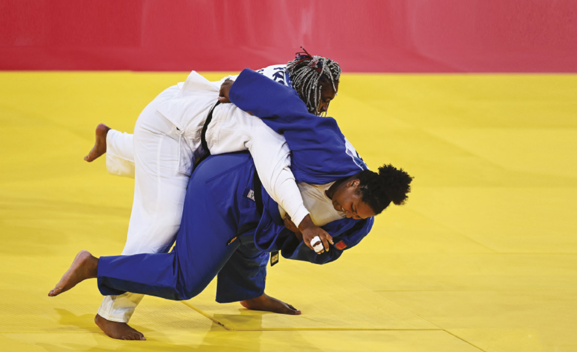 Fotografia de Beatriz Souza, uma mulher de cabelos cacheados presos em um rabo-de-cavalo, vestida com um quimono, executando um movimento de judô em que se joga no chão carregando a adversária consigo, nas costas, passando uma rasteira na perna dela com o pé direito, apoiada somente com o pé esquerdo no chão.