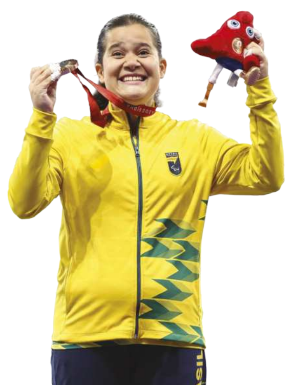 Fotografia de Verônica Hipólito. Ela é uma mulher com cabelos presos em um rabo-de-cavalo, usando um uniforme da equipe olímpica brasileira, sorrindo, com uma medalha de bronze na mão, amarrada ao pescoço. Na outra mão, ela segura um boneco do mascote olímpico de Paris 2024.