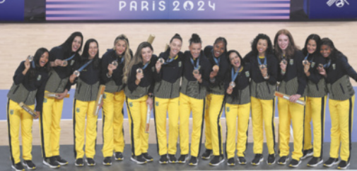 Fotografia de um grupo de jogadoras de vôlei enfileiradas, vestidas com casacos, segurando medalhas penduradas em volta do pescoço.