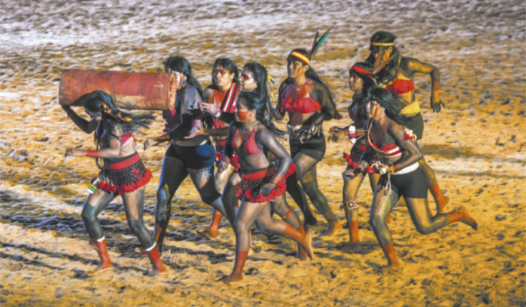 Fotografia de um grupo de mulheres indígenas correndo em uma praia, usando adereços tradicionais indígenas e pinturas corporais. Uma delas está à frente carregando uma tora nas costas.