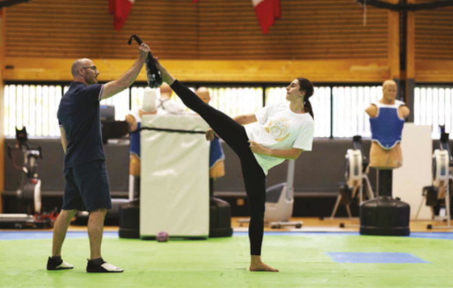 Fotografia de Marzieh Hamidi. Ela é uma mulher de cabelos longos presos em um rabo-de-cavalo, chutando um anteparo segurado por um homem diante dela, à altura da cabeça, em um treinamento em uma academia.