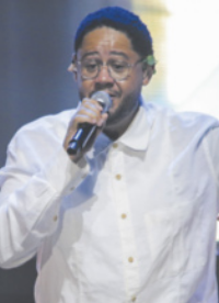 Fotografia de Emicida. Ele é um homem de cabelos curtos e cacheados, usando óculos e uma camisa, cantando com um microfone na mão.