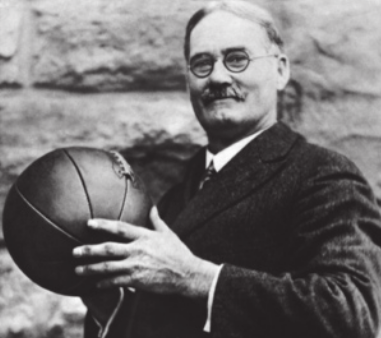 Fotografia em preto e branco de James Naismith. Ele é um homem com bigode e cabelos curtos grisalhos, usando óculos e terno, segurando uma bola de basquete.