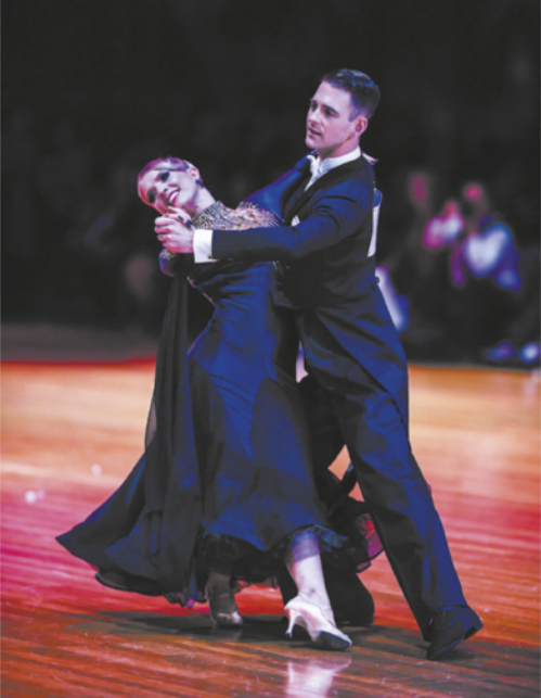Fotografia de um casal dançando de mãos dadas em um palco. O homem, de terno, segura a mão e a cintura da mulher, que está de vestido e salto alto. Ela inclina o corpo, deitando-se no braço dele e se apoiando com o braço ao redor do ombro do parceiro.