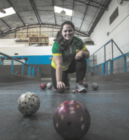 Fotografia de Ana Caroline Martins. Ela é uma mulher de cabelos longos, presos em um rabo-de-cavalo, ajoelhada em uma quadra de bocha, segurando uma bola apoiada no chão. Ao lado dela há pequenas bolas de diversos tamanhos, e à frente dela há duas bolas maiores.