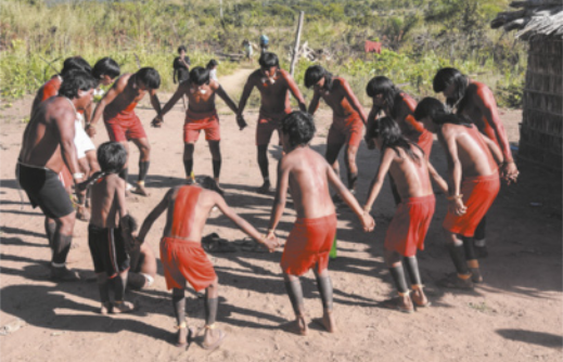 Fotografia de um grupo de indígenas em roda, de mãos dadas, dançando com os joelhos levemente flexionados e o tronco inclinado para a frente. Eles dançam sobre um piso de areia, com vegetação ao fundo.