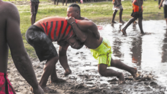 Fotografia de dois homens lutando sobre a areia, próximos ao chão, agarrando-se mutuamente com os braços. Eles estão com os corpos inclinados para a frente, apoiados sobre as pernas flexionadas, tentando derrubar o oponente.