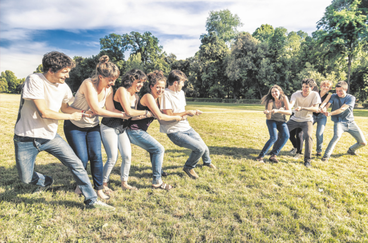 Fotografia de homens e mulheres jovens brincando de cabo de guerra em uma área gramada, com árvores ao fundo. Há 5 pessoas puxando de um lado de uma corda e 4 pessoas puxando do outro lado da corda.