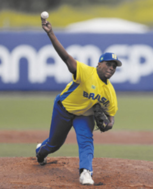 Fotografia de Felipe Natel em um campo de beisebol. Ele é um homem de boné, usando um uniforme da seleção brasileira de beisebol. Ele está arremessando uma bola, com o braço erguido, inclinando o tronco para a frente.