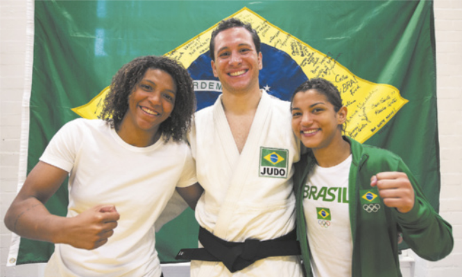 Fotografia de três atletas à frente de um a bandeira do Brasil com autógrafos. À esquerda está Rafaela Silva: uma mulher de cabelos cacheados sorrindo com o punho fechado à frente do corpo; ao centro está Tiago Camilo: um homem sorridente, com cabelos curtos e um quimono; e Sarah Menezes: uma mulher sorrindo com uma jaqueta da seleção brasileira de judô, mostrando o punho fechado à frente do corpo.