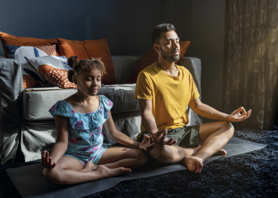 Fotografia de um homem e uma menina praticando meditação, sentados sobre um colchonete estendido no chão de uma sala de estar. Eles estão sentados, de olhos fechados, com as pernas cruzadas, a coluna ereta e as mãos apoiadas sobre os joelhos, unindo os dedos polegares e indicadores em forma de pinça, voltados para cima.