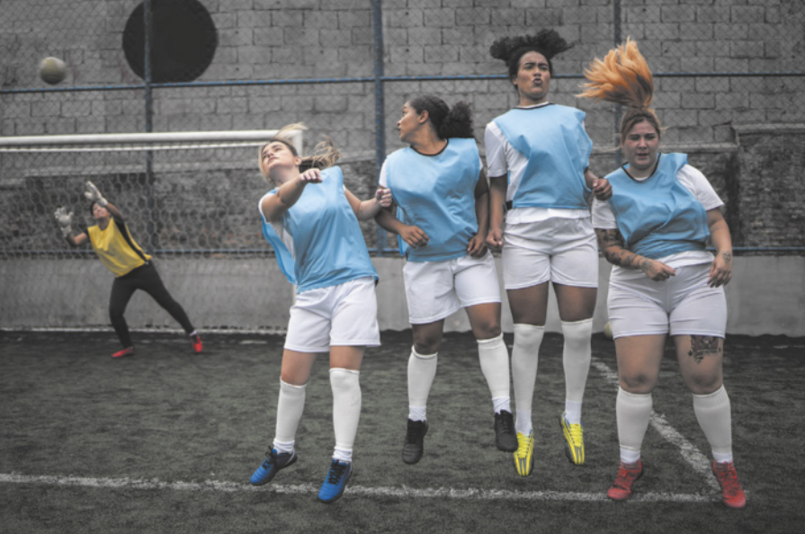 Fotografia de mulheres jogando futebol em uma quadra. Elas estão enfileiradas, usando jalecos, saltando para tentar bloquear a cobrança de falta de uma adversária, com a bola voando em direção ao gol. A goleira se prepara para saltar e defender o gol, usando luvas na mão e um jaleco.