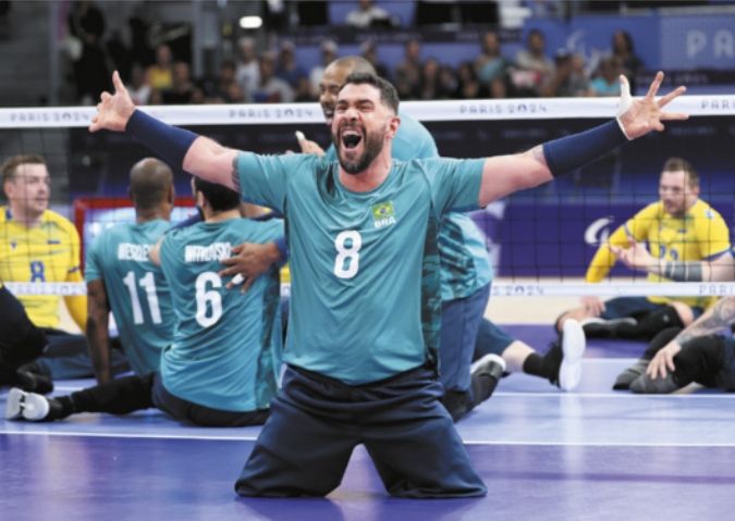 Fotografia de Daniel Yoshizawa. Ele é um homem com barba e cabelos curtos, ajoelhado no chão de uma quadra de vôlei sentado, sorrindo, com os braços abertos em gesto de comemoração.