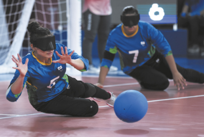Fotografia de Moniza Aparecida de Lima. Ela é uma paratleta com uma máscara sobre os olhos e o nariz, saltando em direção a uma bola, em frente ao gol de uma quadra de golbol. Ela está com o corpo deitado próximo ao chão e as mãos abertas em frente ao rosto. Ao lado dela, está outra paratleta da mesma equipe, ajoelhada no chão, movendo-se em direção à colega de equipe.