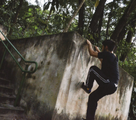 Fotografia de um jovem de tênis e calças de ginástica escalando um muro de concreto ao lado de uma escadaria, com as mãos agarradas ao topo do muro e as pontas dos pés sobre a parede do muro.