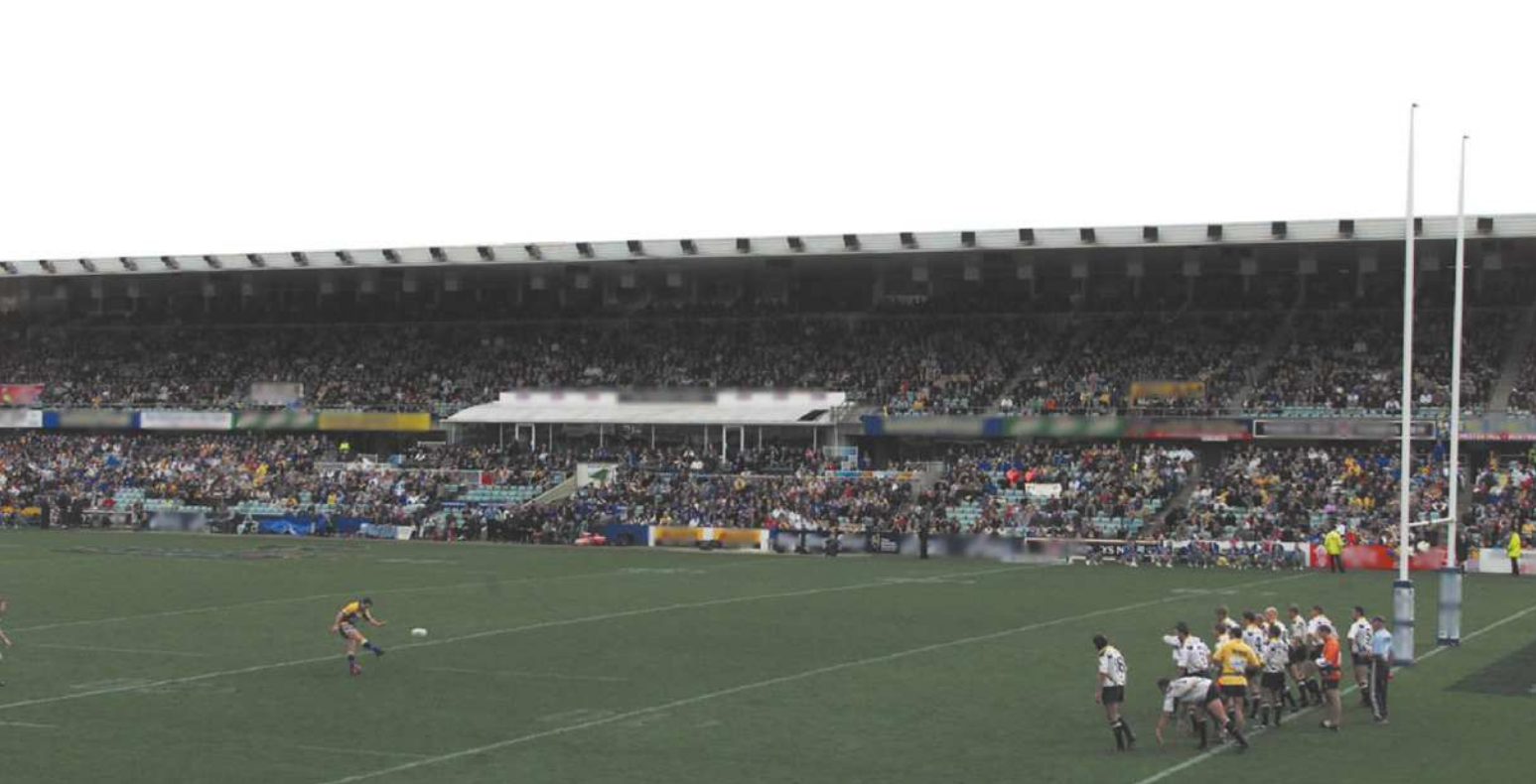 Fotografia de uma partida de rúgbi em um estádio com um campo de grama e arquibancadas. Um jogador está chutando uma bola, e a alguns metros dele estão enfileirados um grupo de jogadores adversários, formando uma linha defensiva.
