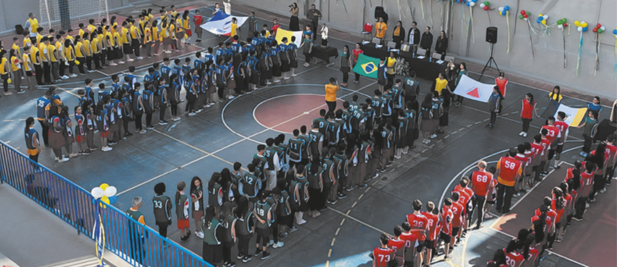 Fotografia de estudantes em filas em uma quadra poliesportiva. As filas de estudantes, identificados com jalecos de cores diferentes, estão em pé diante de bandeiras diversas e de uma mesa com jurados, que estão em pé atrás da mesa.