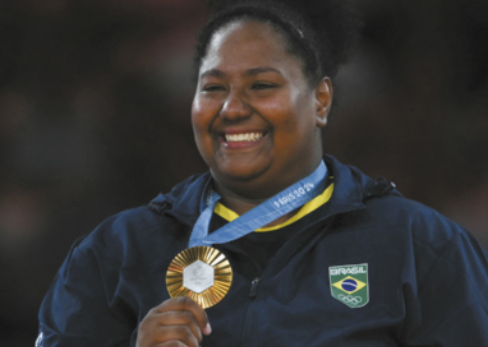 Fotografia de Beatriz Souza. Ela é uma mulher de cabelos cacheados, usando uma tiara, segurando uma medalha de ouro pendurada ao pescoço.