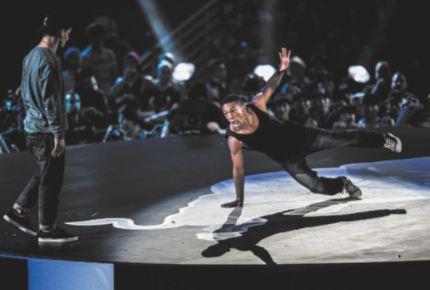 Fotografia de um homem dançando breaking em um palco, com o corpo deitado no ar, apoiado sobre a palma de uma das mãos e a ponta de um dos pés, erguendo a outra perna e o outro braço em diagonal, equilibrando-se nessa posição. Diante dele está outro dançarino em pé, que o observa. Ao fundo, uma plateia assiste à batalha.