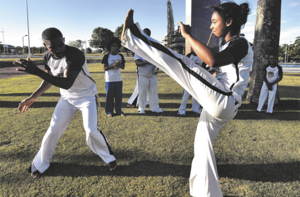 Fotografia de uma garota e um homem em uma roda de capoeira no gramado de uma praça. A garota está chutando com a perna estendida para o alto enquanto o homem ginga diante dela, se movimentando lateralmente, com o tronco inclinado e um dos braços à frente do corpo. Ao fundo estão pessoas em pé batendo palmas, cantando e tocando instrumentos como pandeiro e berimbau.