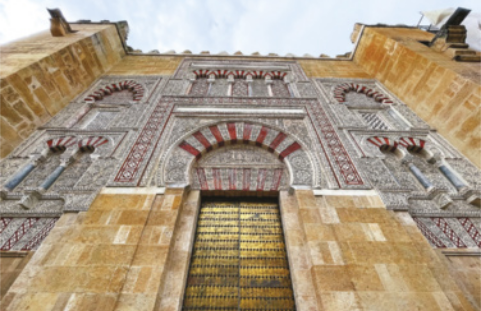 Imagem de uma mesquita. Ela possui duas torres, uma porta central e janelas arqueadas, distribuídas ao longo de três níveis. A porta e as janelas são emolduradas por arcos listrados.