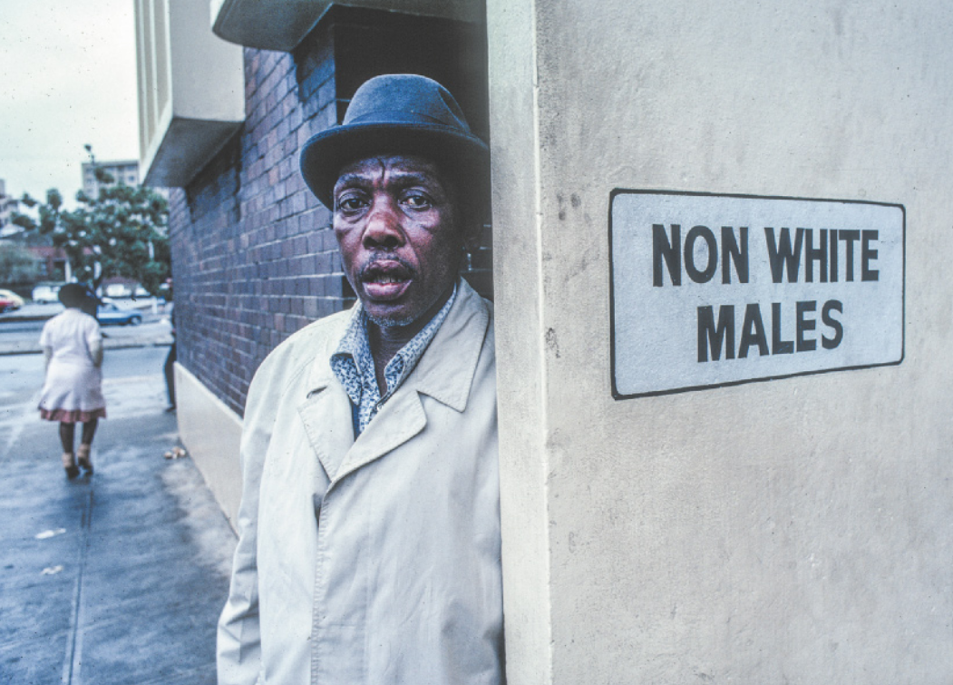 Imagem de homem negro ao lado de uma porta com uma inscrição em inglês. Ele usa chapéu, camisa e casaco.