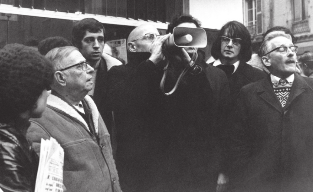 Fotografia em preto e branco de um grupo de pessoas em um protesto. Ao centro, há um homem careca utilizando um megafone. Ao lado dele, à esquerda, encontra-se um indivíduo mais baixo, idoso, usando óculos e jaqueta.