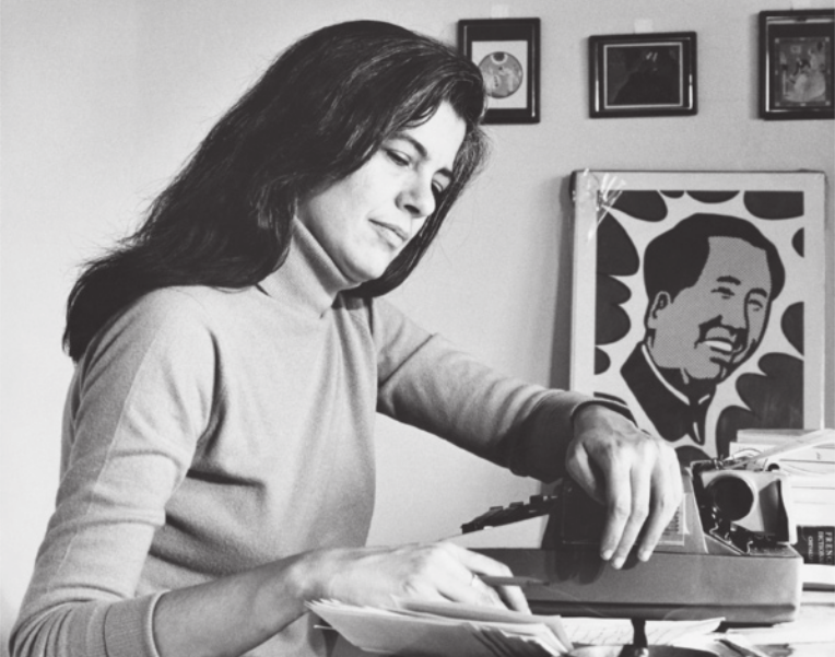 Fotografia em preto e branco de Susan Sontag. Ela está diante de uma mesa, com o braço esquerdo apoiado sobre uma máquina de escrever e a mão direita folheando um caderno.