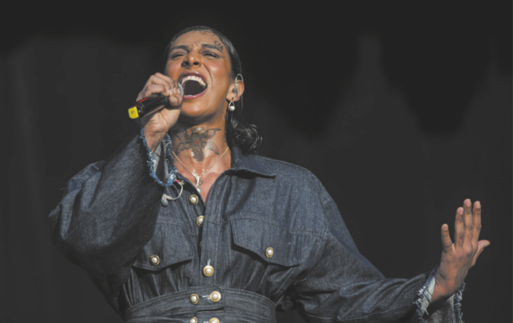 Fotografia de Linn da Quebrada cantando com os olhos fechados. Ela é negra, possui tatuagens no rosto e no pescoço, está com cabelo preso, usa camisa com botões perolados e utiliza um microfone.