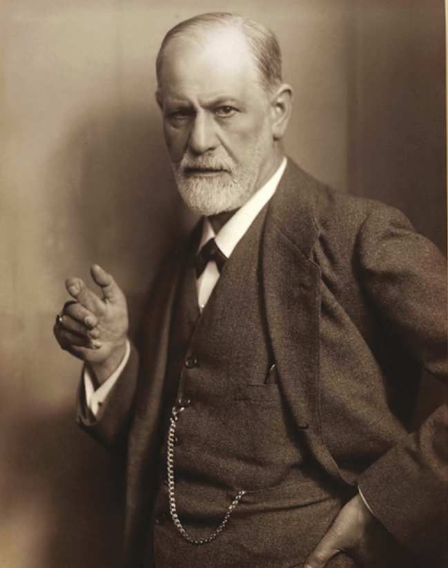 Fotografia em preto e branco de Sigmund Freud. Ele é branco, possui cabelo e barba curtos, usa camisa, colete e paletó e está com a mão direita levantada, próxima ao peito.