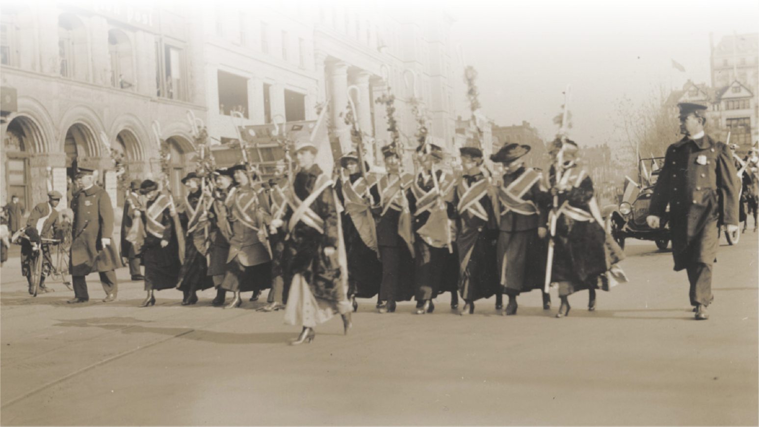Imagem em preto e branco de mulheres. Elas usam chapéu, casaco, saia longa e duas faixas cruzadas no peito, carregam uma haste com o topo curvado e caminham lado a lado em uma rua. À frente delas, encontra-se uma mulher com a mesma vestimenta. Dois policiais acompanham o grupo.