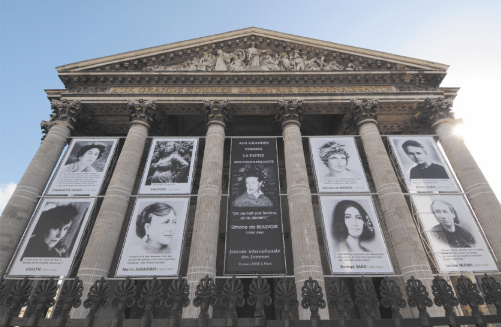 Imagem de painéis pendurados na fachada de um edifício. Os painéis contêm nove retratos femininos em preto e branco. O edifício possui seis colunas e um frontão com esculturas.