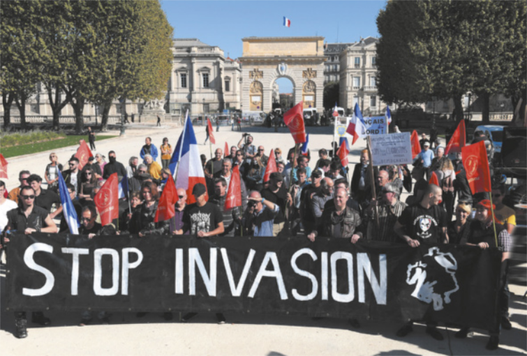 Imagem de grupo de manifestantes em uma praça arborizada. Os indivíduos à frente do grupo carregam um cartaz com uma frase em inglês. Os demais manifestantes carregam outros cartazes e bandeiras.