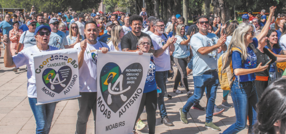 Imagem de manifestantes caminhando em uma praça, em um dia ensolarado. Eles carregam cartazes com um coração que contém a letra A e a bandeira do Brasil.
