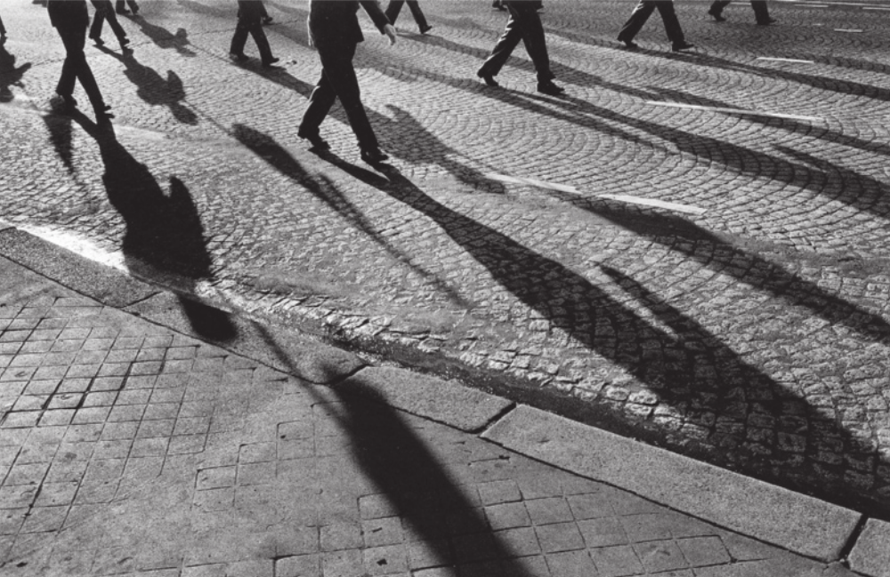 Imagem de pessoas caminhando em uma rua, na mesma direção. A sombra delas está projetada no chão.
