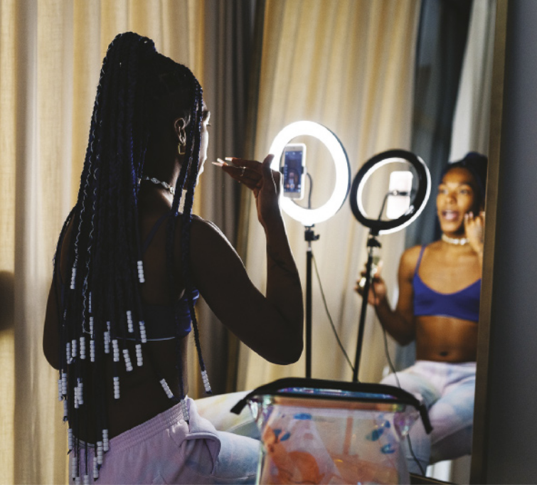 Imagem de mulher negra jovem sentada, com um smartphone posicionado na altura do rosto e preso no centro de um anel luminoso. Ela usa calça e top, possui unhas longas e tranças no cabelo e gesticula com a mão direita.