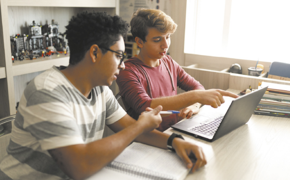 Imagem de dois garotos estudando. Eles estão sentados diante de uma mesa e utilizam notebook, caderno e livros.