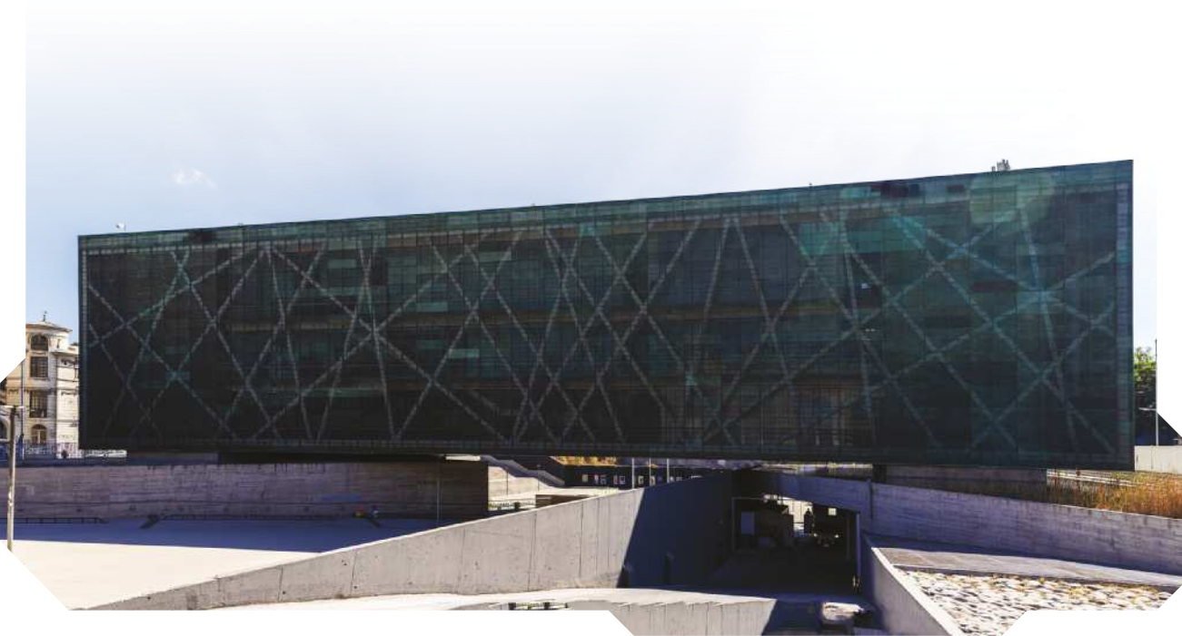 Imagem de edifício retangular, com comprimento maior do que a altura. A fachada contém diversas janelas de vidro e há um túnel abaixo do edifício.