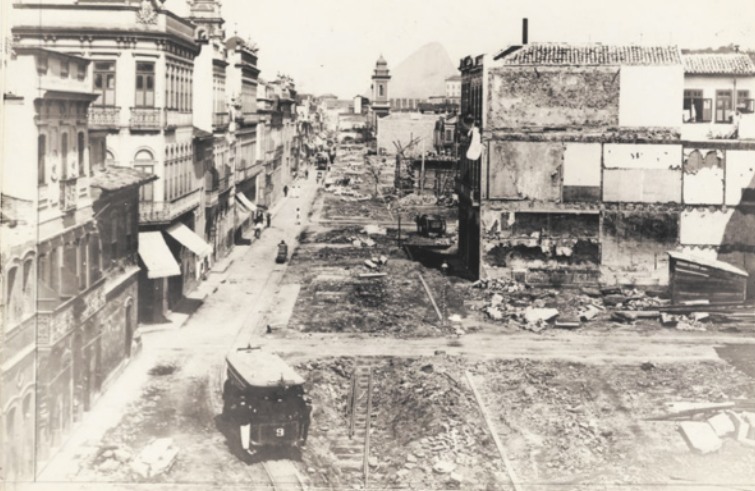Imagem em preto e branco de uma rua com trilhos. À esquerda dos trilhos, a rua é pavimentada e cercada por edifícios de até três pavimentos. A área à direita dos trilhos é de terra, com alguns edifícios demolidos. Um bonde e pedestres transitam pela rua.