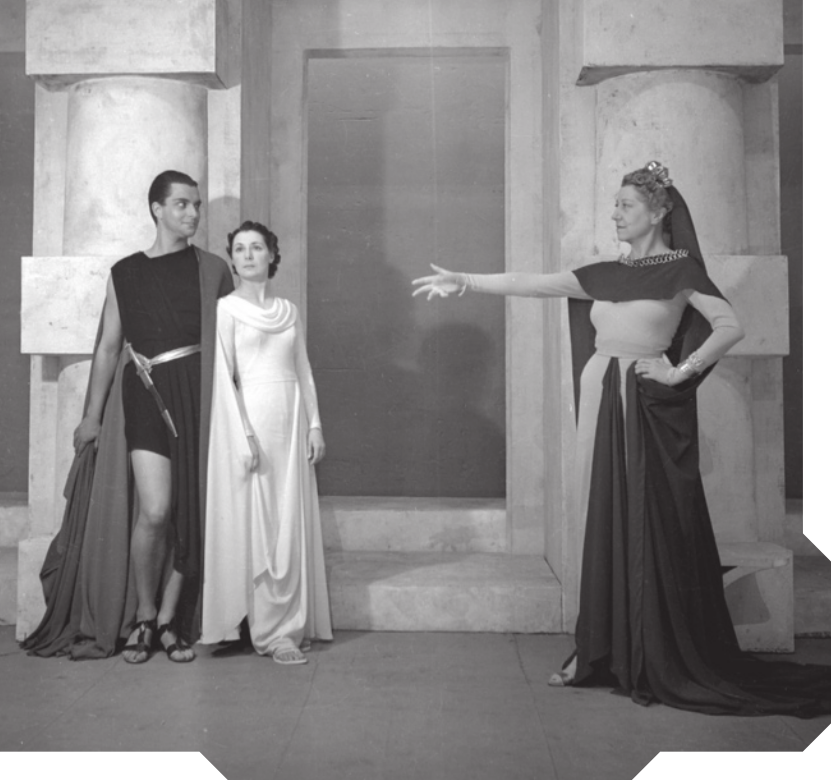 Imagem da cena de uma peça de teatro. Um dos personagens, à direita, é uma mulher que está com a mão esquerda na cintura e a mão direita estendida para frente. À esquerda, um homem e uma mulher estão lado a lado, encostados em uma das colunas do cenário.