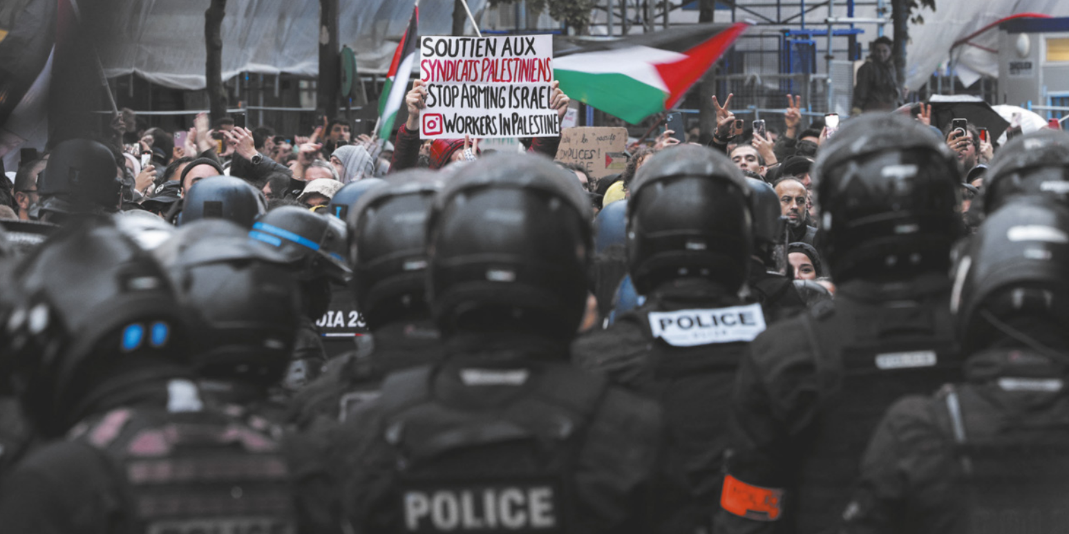 Imagem de policiais avançando em direção a manifestantes, que seguram bandeiras da Palestina e um cartaz.