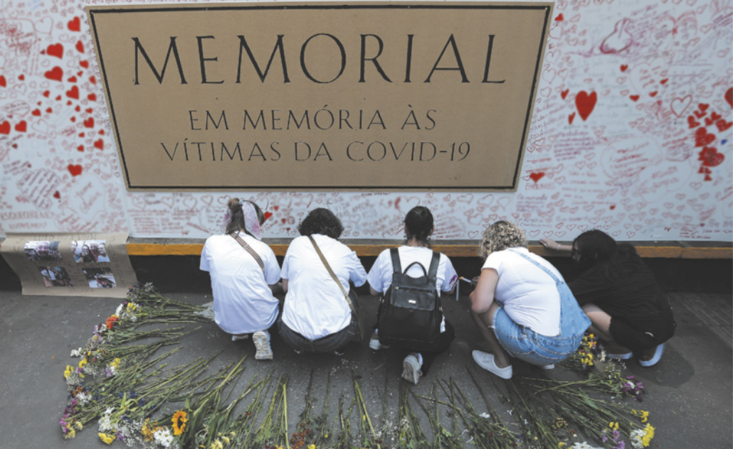 Imagem de cinco pessoas agachadas em frente a uma placa, que contém as seguinte identificação: Memorial em memória às vítimas da Covid-19. Na parede, ao redor da placa, há inscrições e corações desenhados. Em torno das pessoas, há flores dispostas em semicírculo no chão.