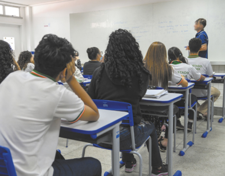 Imagem de sala de aula. O professor está em frente ao quadro branco e os estudantes estão sentados diante de mesas, organizadas em fileiras.