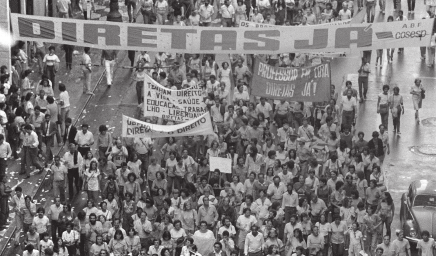 Imagem em preto e branco de manifestantes caminhando em uma rua. Eles carregam faixas, uma delas contém a expressão Diretas Já.