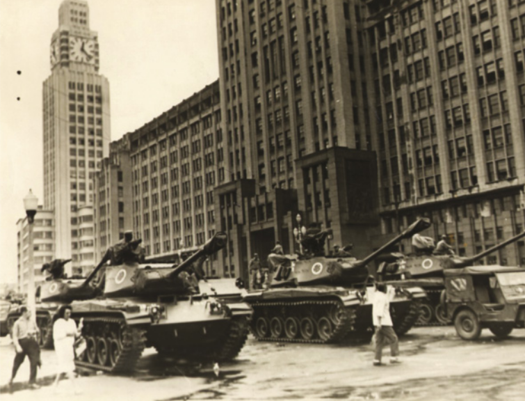 Imagem em preto e branco de tanques de guerra. Eles estão em uma via cercada por edifícios.