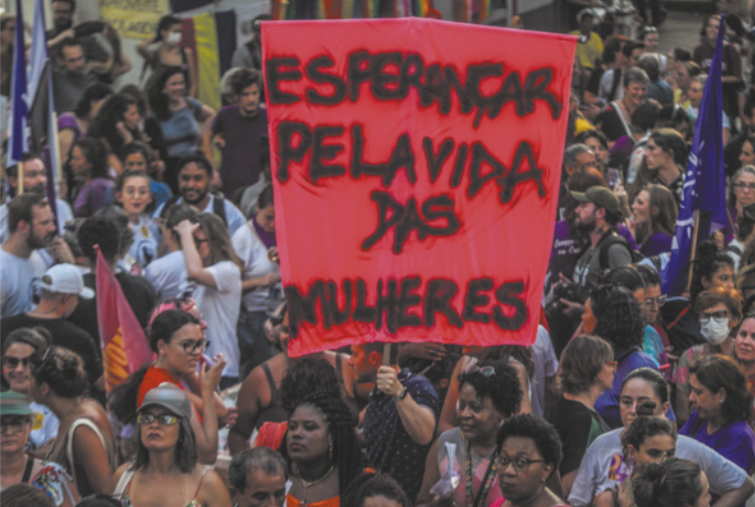 Imagem de manifestantes com bandeiras e um cartaz, no qual está escrito: esperançar pela vida das mulheres.