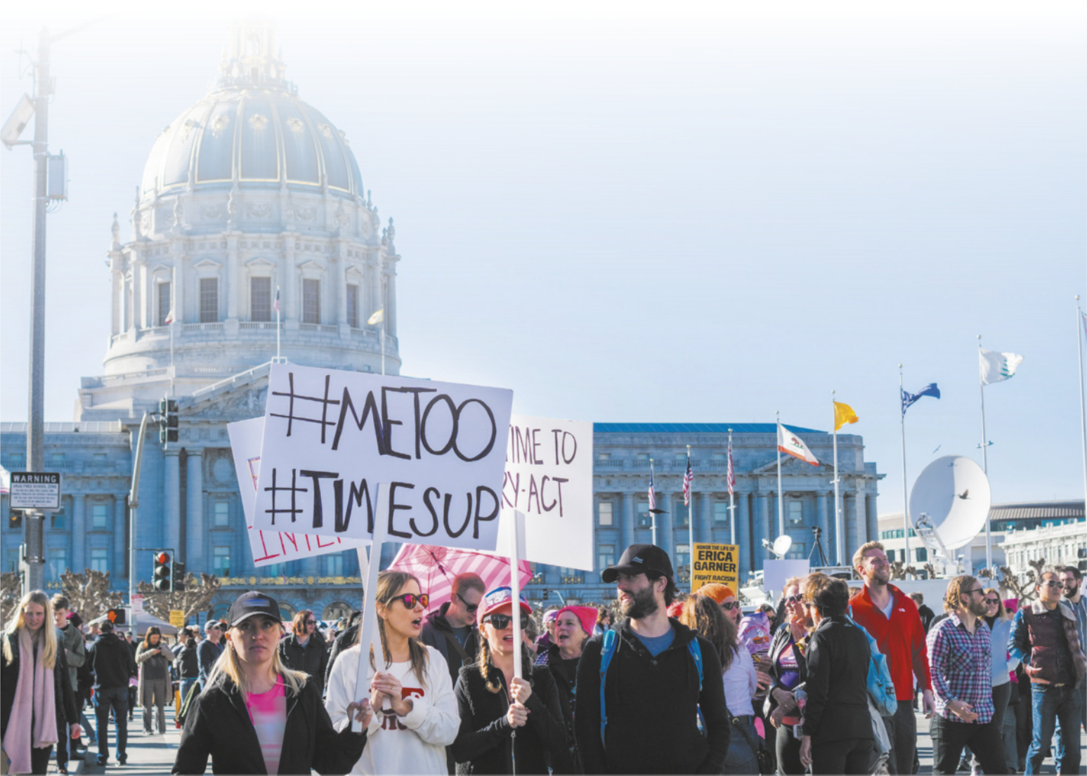 Imagem de manifestantes em uma praça. Três mulheres carregam placas com frases em inglês e hashtags.