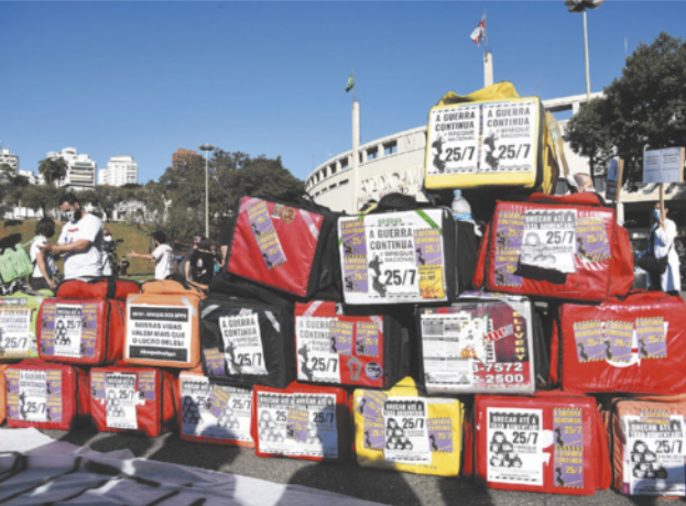 Imagem de cartazes colados em mochilas térmicas, que estão no chão de uma praça.