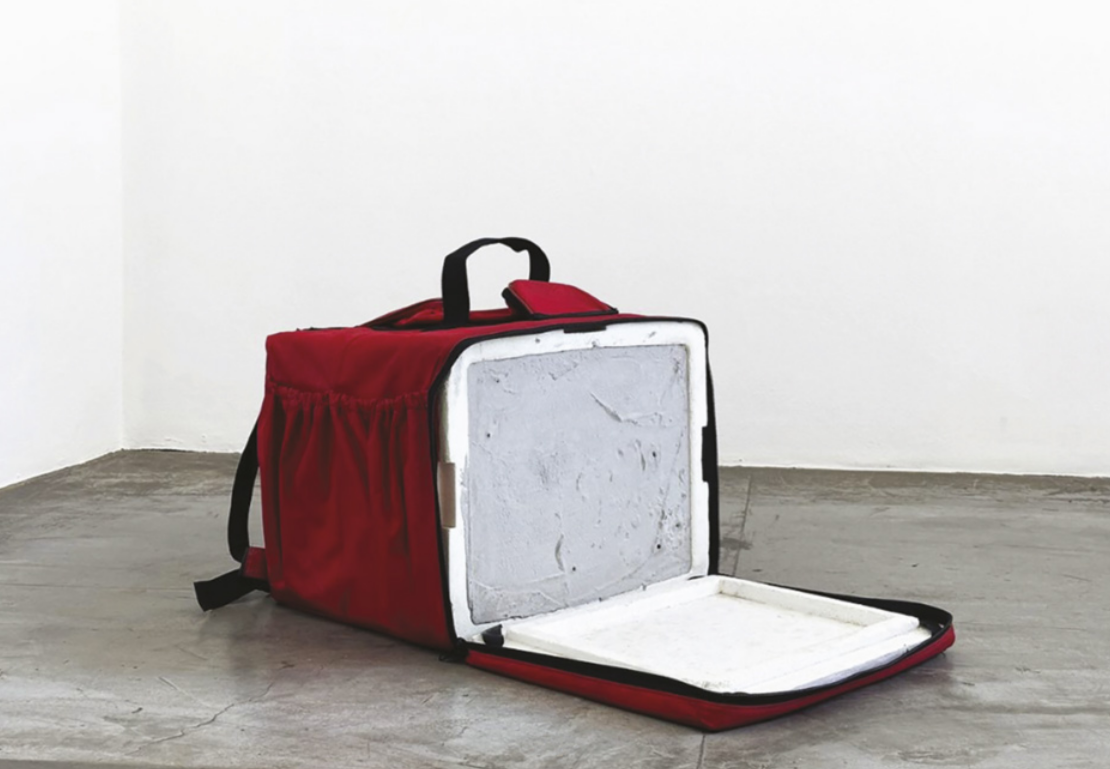 Imagem de mochila térmica no piso de uma sala. A mochila é revestida com isopor e está cheia de cimento.