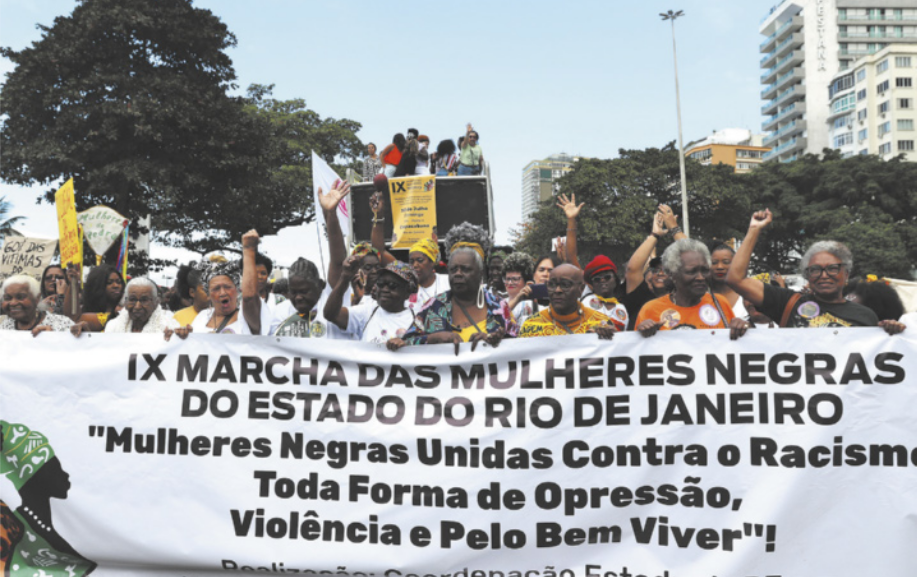 Imagem de mulheres negras caminhando em uma rua. Elas carregam uma faixa com a seguinte frase: Mulheres negras unidas contra o racismo, toda forma de opressão, violência e pelo bem viver.